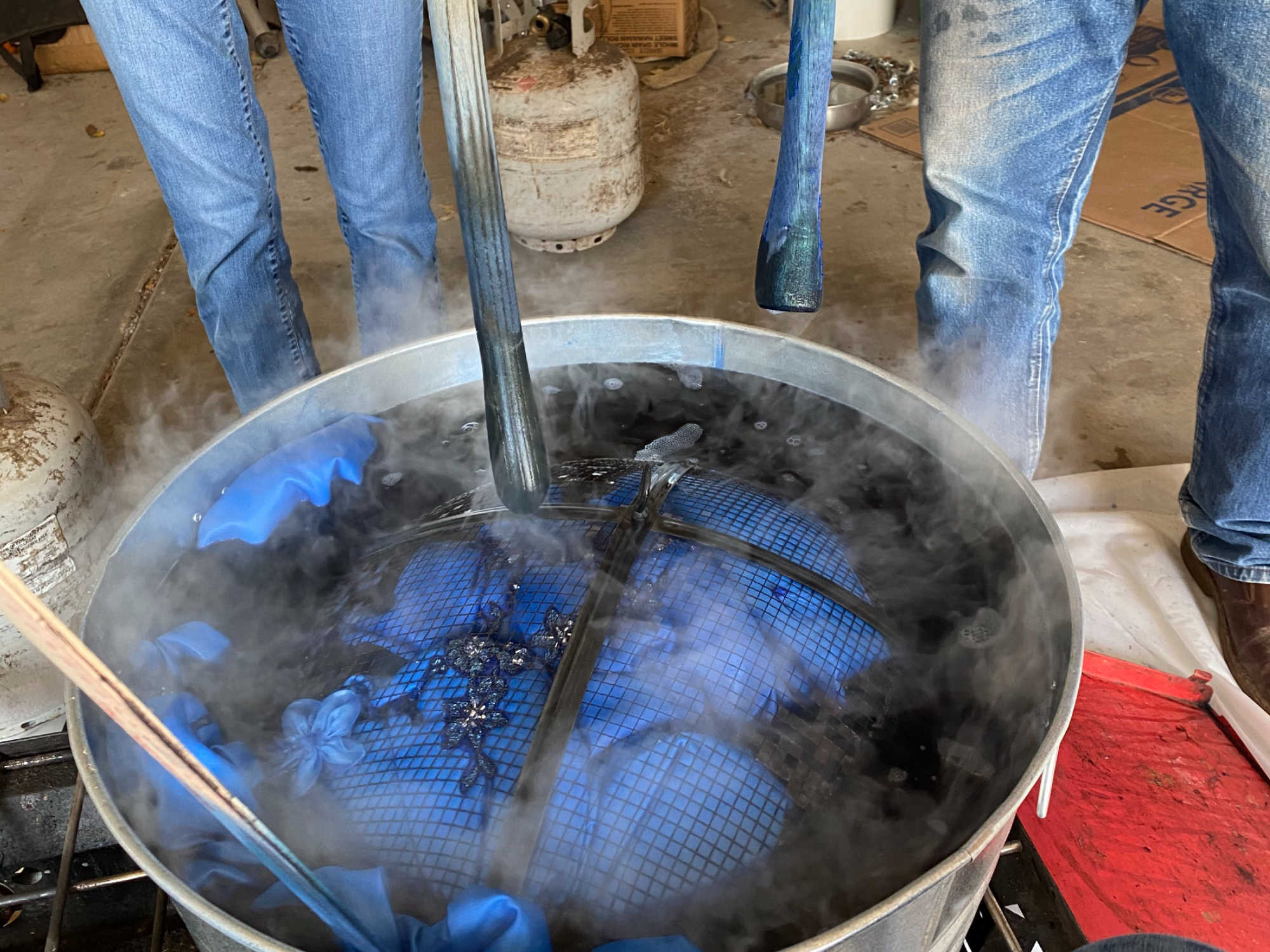 A secondhand wedding dress being dyed blue in a metal tub. Several people use yardsticks and a fire pit cover to hold the dress underwater.